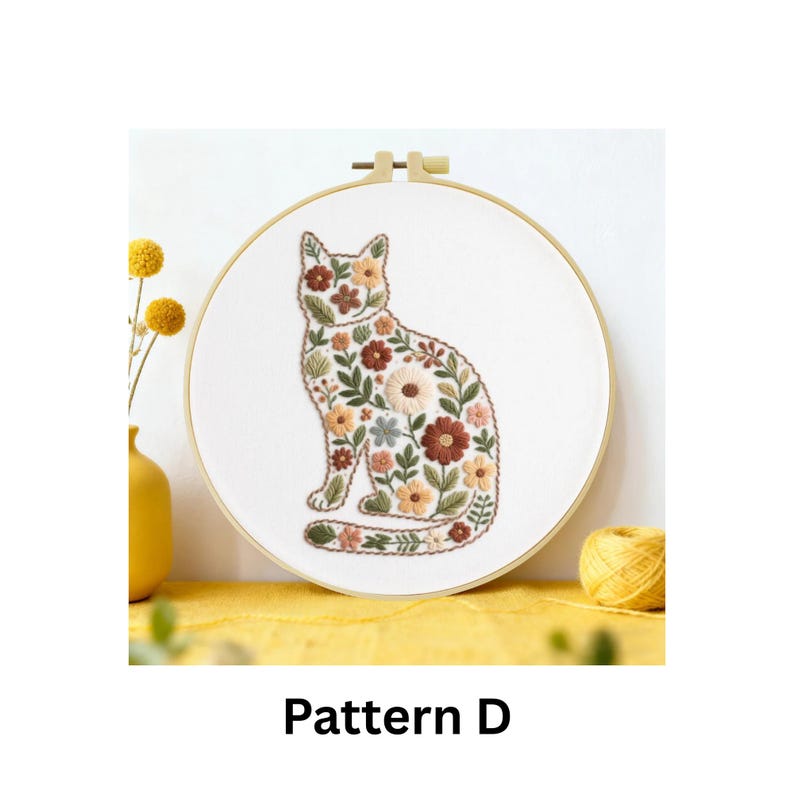 Puede incluir: Un dise&ntilde;o de gato bordado con flores sobre tela blanca, dentro de un bastidor de madera. El gato est&aacute; relleno de flores marrones, naranjas y verdes. El texto "Pattern D" est&aacute; en la parte inferior.