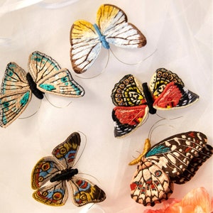 May include: Five embroidered butterflies in various colors and patterns, including blue, red, yellow, and black, on a white background.