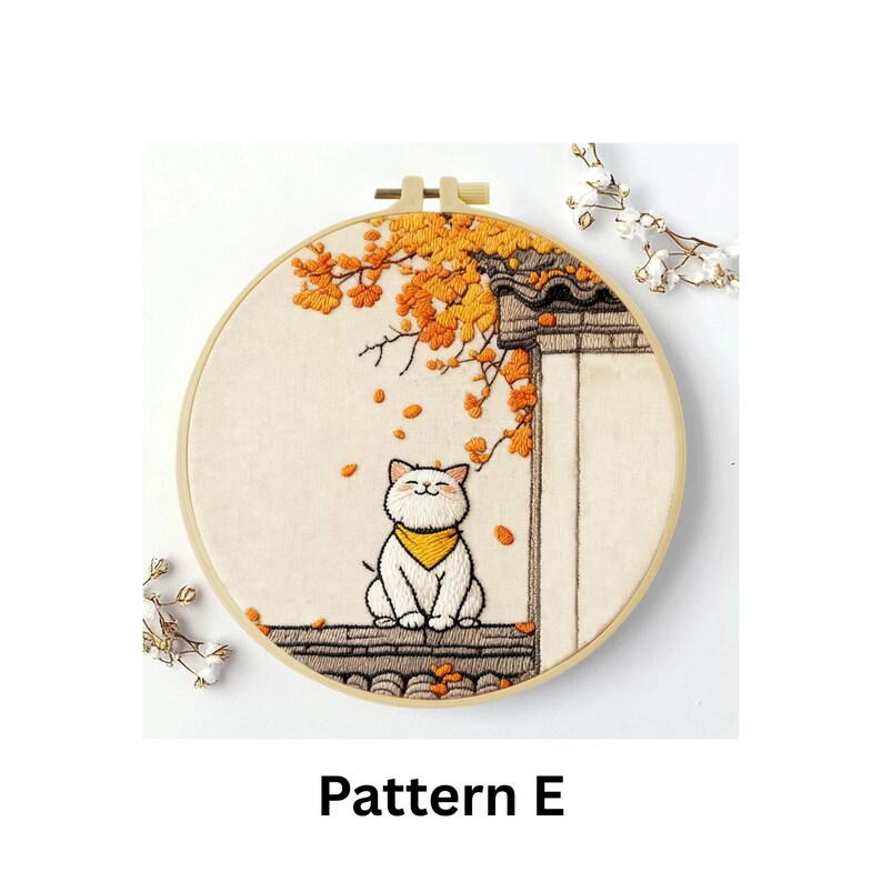 Puede incluir: Arte de aro bordado que presenta un gato blanco sonriente con una bufanda amarilla, posado en un tejado. La escena incluye follaje oto&ntilde;al y un edificio tradicional. El texto "Pattern E" se muestra en la parte inferior.