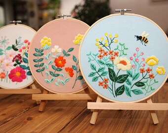 May include: Three embroidery hoops with floral designs. The hoops are on wooden stands and feature different colored fabric with embroidered flowers, leaves, and a bee.