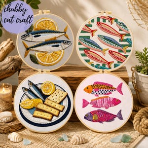 May include: Four circular embroidery hoops featuring fish-themed designs. One shows two fish with lemon slices, another has a pattern of small fish on green stripes, a third depicts fish with crackers on a plate, and the fourth shows three colourful fish.
