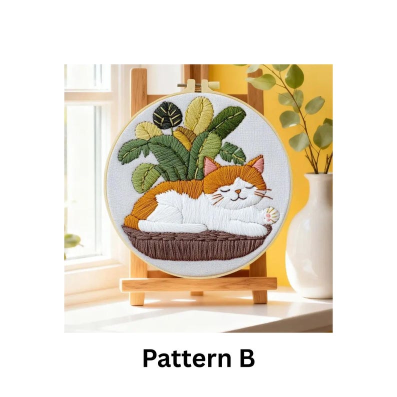 Puede incluir: Arte de aro bordado con un gato naranja y blanco durmiendo en una cesta tejida con follaje verde y amarillo. El bordado se exhibe en un caballete de madera. El texto "Pattern B" est&aacute; en la parte inferior.