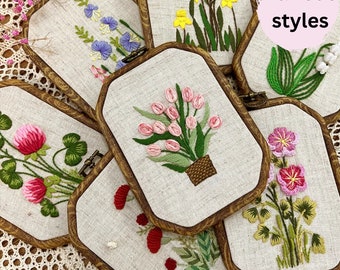 May include: Six embroidery hoops with various floral designs, including pink tulips, clover, and lilies. The hoops are made of wood and have a natural finish. The text "Various styles" is written on a pink background in the upper right corner of the image.