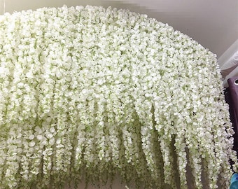 May include: A white artificial wisteria garland with long, cascading strands of delicate white flowers. The garland is perfect for adding a touch of elegance and beauty to any space.
