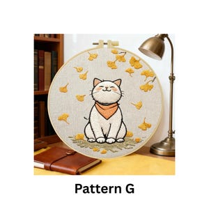 Puede incluir: Arte de aro de gato bordado con un gato blanco sonriente con un pa&ntilde;uelo naranja, rodeado de hojas de ginkgo amarillas. El dise&ntilde;o est&aacute; sobre una tela beige, en un aro de madera. El texto "Pattern G" est&aacute; en la parte inferior.