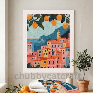 May include: A framed art print showing a vibrant coastal town. The artwork features buildings in shades of pink, orange, and yellow, with a blue sea and mountains. Orange tree branches are at the top. The text "chubbycatcraft" is at the bottom of the image.