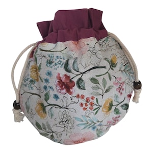 May include: A round drawstring pouch with a floral pattern in shades of red, pink, yellow, and blue on a cream background. The top is a solid burgundy fabric, and the drawstring is a natural rope with wooden beads.