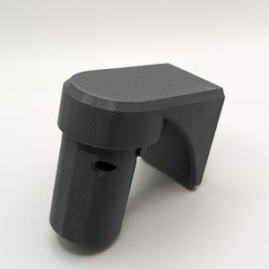 Op de afbeelding: Een 3D-geprint zwart kunststof onderdeel met een afgeronde bovenkant en een gat aan de zijkant. Het onderdeel is ontworpen om aan een muur of ander oppervlak te worden bevestigd.