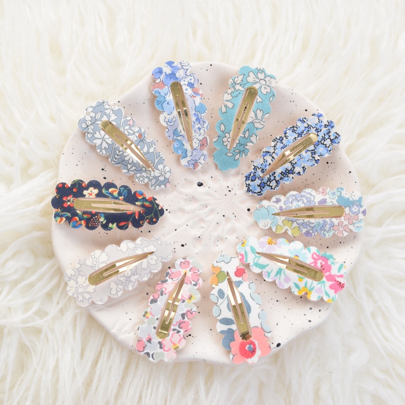 May include: A set of 10 floral patterned hair clips with scalloped edges. The clips are arranged in a circle on a white plate with black speckles. The clips are in various colors, including blue, pink, and green.