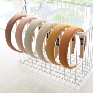 May include: A set of six wide headbands in various neutral colors, including beige, brown, and pink. The headbands are made of a soft, fuzzy material and are displayed in a white wire basket.