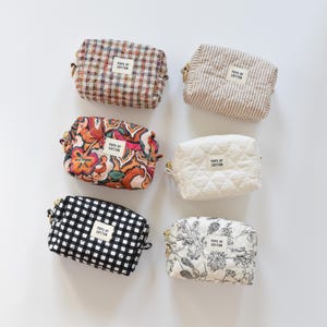 May include: Six small, rectangular cosmetic bags in various patterns and colors. Designs include plaid, stripes, floral, quilted, and gingham. Each bag has a zipper closure and a small label that reads "POPS OF COTTON."