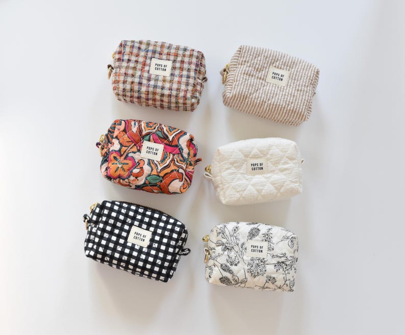 May include: Six small, rectangular cosmetic bags in various patterns and colors. Designs include plaid, stripes, floral, quilted, and gingham. Each bag has a zipper closure and a small label that reads "POPS OF COTTON."
