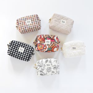 May include: A collection of six small cosmetic bags in various patterns and colors. Designs include gingham, floral, striped, and quilted textures. Each bag has a gold zipper and a label that reads "POPS OF COTTON".