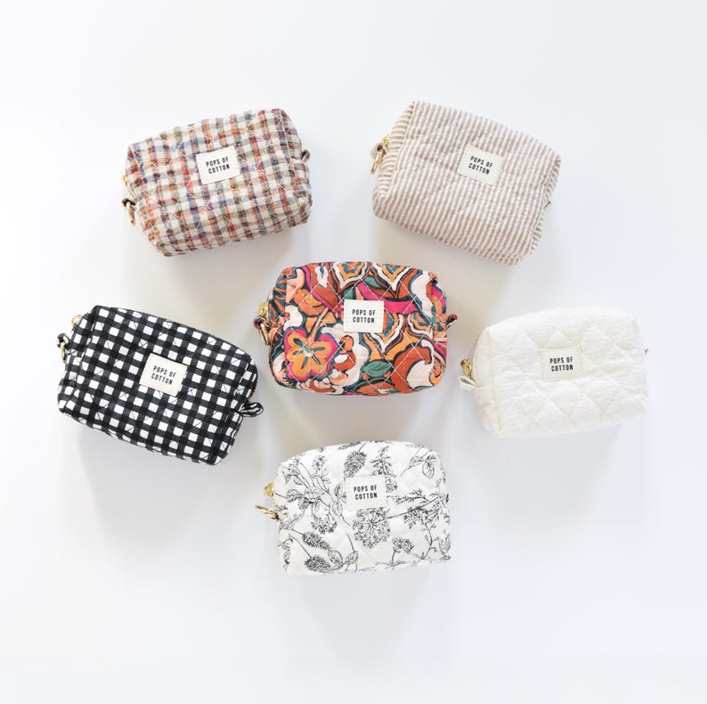 May include: A collection of six small cosmetic bags in various patterns and colors. Designs include gingham, floral, striped, and quilted textures. Each bag has a gold zipper and a label that reads "POPS OF COTTON".