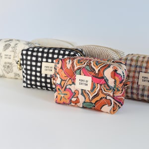 May include: A collection of small, quilted cosmetic bags in various patterns. Designs include floral, black and white checkered, abstract floral, and plaid. Each bag has a zipper closure and a label that reads "Pops of Cotton."