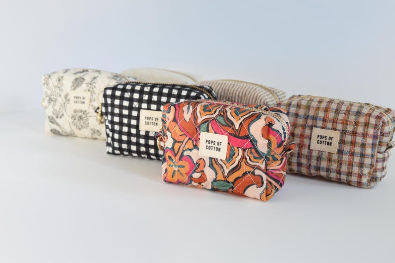 May include: A collection of small, quilted cosmetic bags in various patterns. Designs include floral, black and white checkered, abstract floral, and plaid. Each bag has a zipper closure and a label that reads "Pops of Cotton."