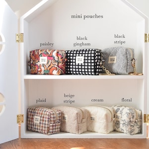 May include: A white house-shaped cabinet displays several mini pouches. The pouches feature various patterns, including paisley, black gingham, black stripe, plaid, beige stripe, cream, and floral. Each pouch has a small label.
