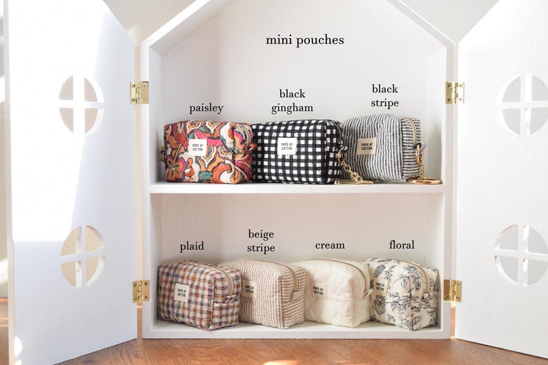 May include: A white house-shaped cabinet displays several mini pouches. The pouches feature various patterns, including paisley, black gingham, black stripe, plaid, beige stripe, cream, and floral. Each pouch has a small label.