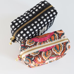May include: Two small, quilted cosmetic bags. One is black and white gingham, the other features a colorful floral pattern in pink, orange, and teal. Both have gold zippers and small gold zipper pulls. The bags are rectangular with rounded ends.