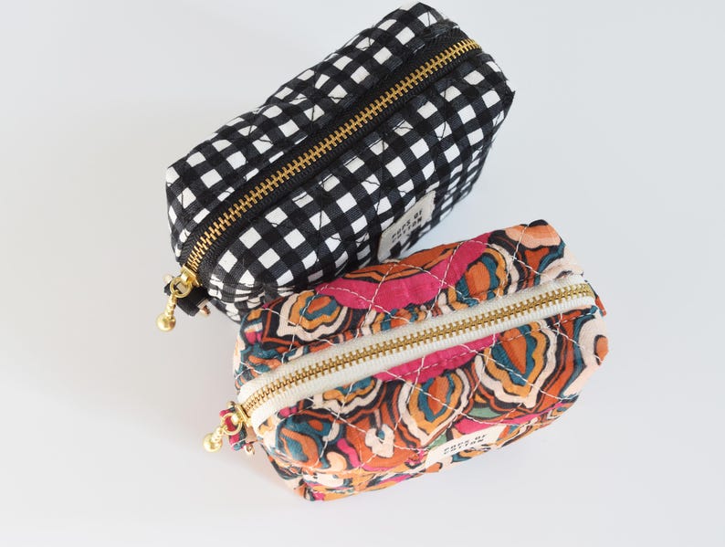 May include: Two small, quilted cosmetic bags. One is black and white gingham, the other features a colorful floral pattern in pink, orange, and teal. Both have gold zippers and small gold zipper pulls. The bags are rectangular with rounded ends.