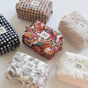 May include: An assortment of quilted cosmetic bags in various patterns and colors, including black and white gingham, floral, and paisley designs. Each bag has a gold zipper and a label that reads "POPS OF COTTON."
