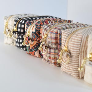 May include: A row of six small, patterned cosmetic bags with gold zippers and matching pulls. The bags feature various designs, including floral, gingham, and plaid patterns. The bags are arranged in a row, with the zippers facing forward.