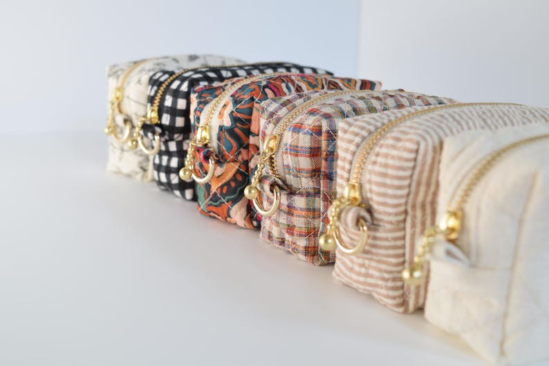 May include: A row of six small, patterned cosmetic bags with gold zippers and matching pulls. The bags feature various designs, including floral, gingham, and plaid patterns. The bags are arranged in a row, with the zippers facing forward.