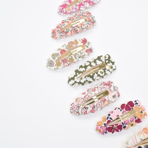 May include: A set of eight floral patterned hair clips with gold metal clasps. The clips are arranged in a row, with the largest clip at the top and the smallest clip at the bottom. The clips are all different colors and patterns, but they all have a floral theme.