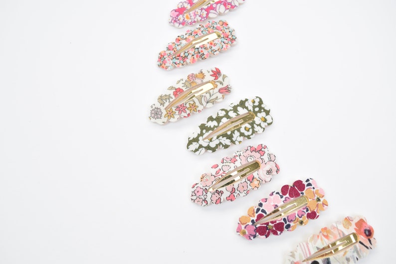 May include: A set of eight floral patterned hair clips with gold metal clasps. The clips are arranged in a row, with the largest clip at the top and the smallest clip at the bottom. The clips are all different colors and patterns, but they all have a floral theme.