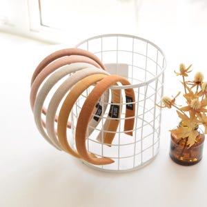 May include: A collection of fabric-covered headbands in various warm tones, including peach, cream, and golden yellow, are displayed in a white wire basket. The headbands have a simple, rounded design, perfect for everyday wear.