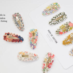 May include: A set of 9 floral patterned hair clips with gold metal clasps. The clips are arranged on a white background with the text "Pops of Cotton" and "Made with Liberty Fabric".