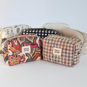 May include: A collection of six small, zippered cosmetic bags in various patterns. The bags feature floral, plaid, and striped designs in colors like orange, black, and cream. Each bag has a small label that reads "POPS OF COTTON".