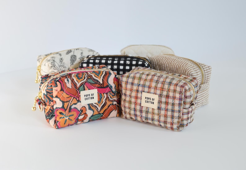 May include: A collection of six small, zippered cosmetic bags in various patterns. The bags feature floral, plaid, and striped designs in colors like orange, black, and cream. Each bag has a small label that reads "POPS OF COTTON".