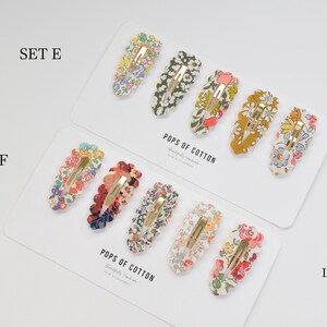 May include: Two sets of hair clips with floral patterns. The clips are made with Liberty fabric and are labeled "Set E" and "Set F". The packaging includes the text "Pops of Cotton" and "Made with Liberty Fabric".