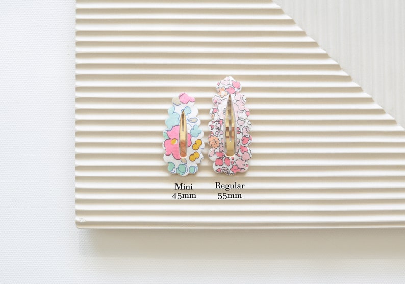 May include: Two floral patterned hair clips, one in pink and one in blue, with gold metal clasps. The smaller clip is labeled "Mini 45mm" and the larger clip is labeled "Regular 55mm".
