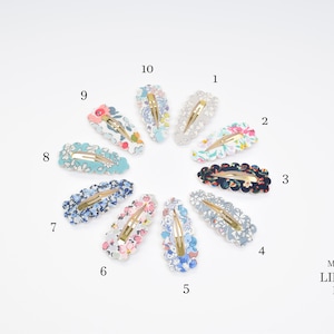 May include: Ten floral patterned hair clips in various colors, including blue, pink, white, and black. The clips are arranged in a circle on a white background. The text "Made with LIBERTY Fabric" is visible in the bottom right corner.
