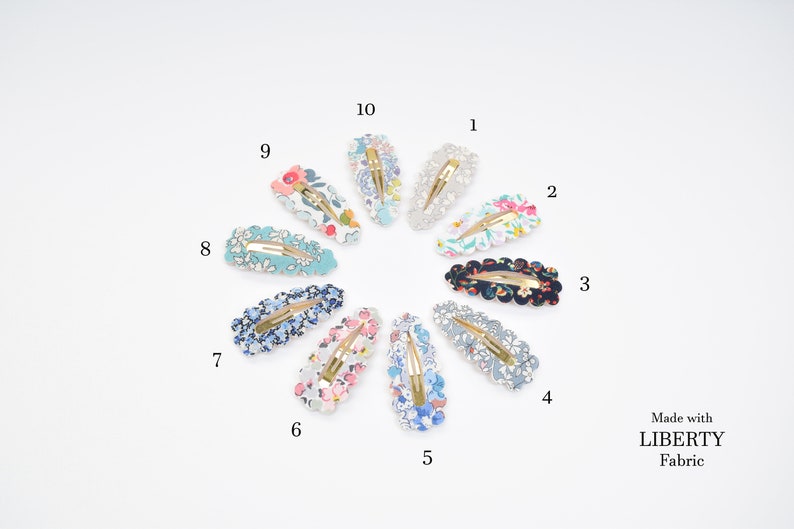 May include: Ten floral patterned hair clips in various colors, including blue, pink, white, and black. The clips are arranged in a circle on a white background. The text "Made with LIBERTY Fabric" is visible in the bottom right corner.
