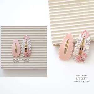 May include: Two pink hair clips with scalloped edges. The larger clip is 5.5 inches and features a floral print fabric. The smaller clip is 4.5 inches and is solid pink. The text "made with LIBERTY fabric & Linen" is printed below the clips.