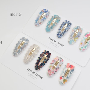 May include: Two sets of hair clips with floral patterns. The top set is labeled "SET G" and the bottom set is labeled "SET H". Both sets are made with Liberty Fabric and are labeled "POPS OF COTTON".