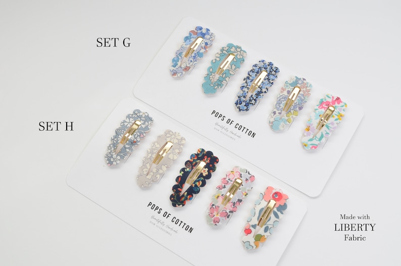 May include: Two sets of hair clips with floral patterns. The top set is labeled "SET G" and the bottom set is labeled "SET H". Both sets are made with Liberty Fabric and are labeled "POPS OF COTTON".