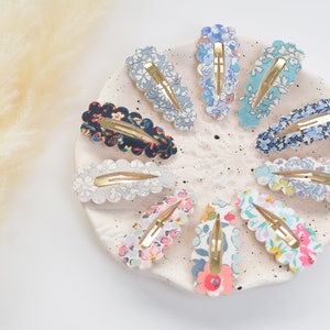 May include: A set of 10 floral patterned hair clips with scalloped edges. The clips are arranged in a circle on a white plate. The clips are in various colors, including blue, pink, and green.