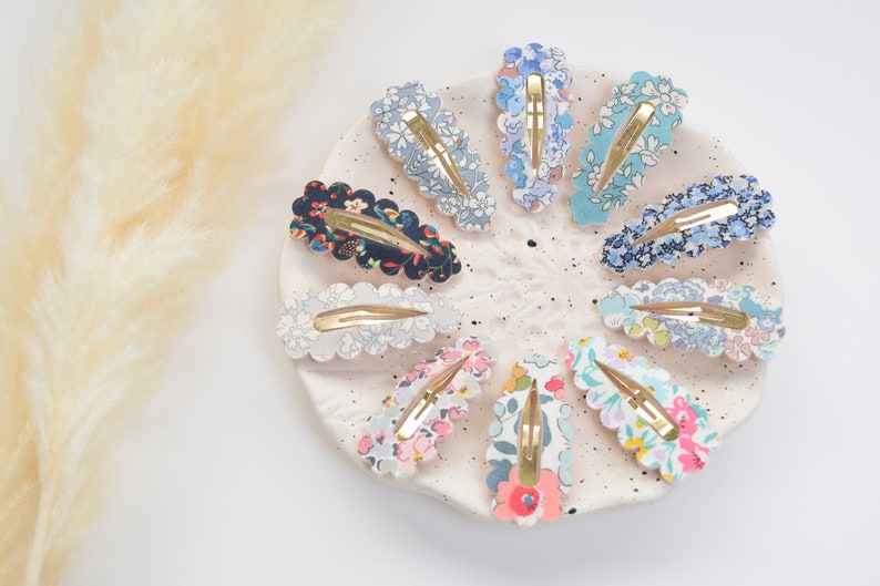 May include: A set of 10 floral patterned hair clips with scalloped edges. The clips are arranged in a circle on a white plate. The clips are in various colors, including blue, pink, and green.