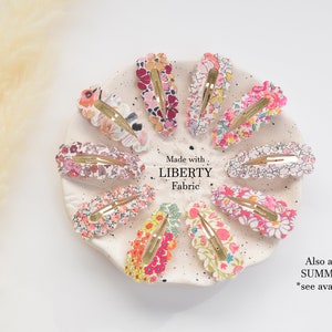 May include: A set of 10 colorful floral hair clips made with Liberty Fabric. The clips are arranged in a circle on a white plate. The text "Made with Liberty Fabric" is printed on the plate. The text "Also available in SUMMER Fabrics *see available listing" is printed below the plate.