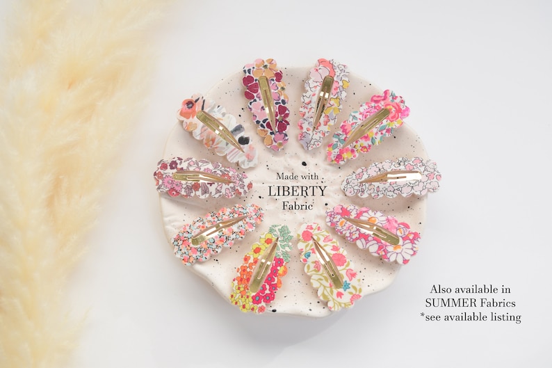 May include: A set of 10 colorful floral hair clips made with Liberty Fabric. The clips are arranged in a circle on a white plate. The text "Made with Liberty Fabric" is printed on the plate. The text "Also available in SUMMER Fabrics *see available listing" is printed below the plate.