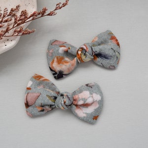 May include: Two floral fabric hair bows in shades of gray, orange, and pink. The bows are tied in a knot and are ready to be worn in hair.