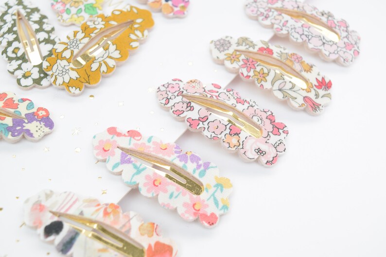 May include: A set of 10 colorful floral patterned hair clips with gold metal clasps. The clips are shaped like a scallop shell and have a white background with pink, yellow, green, and purple flowers.