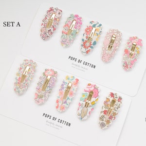 May include: Two sets of hair clips with floral patterns. Set A has three clips with pink, white, and green floral patterns. Set B has three clips with pink, white, and blue floral patterns. The clips are made with Liberty fabric and are labeled "Pops of Cotton".