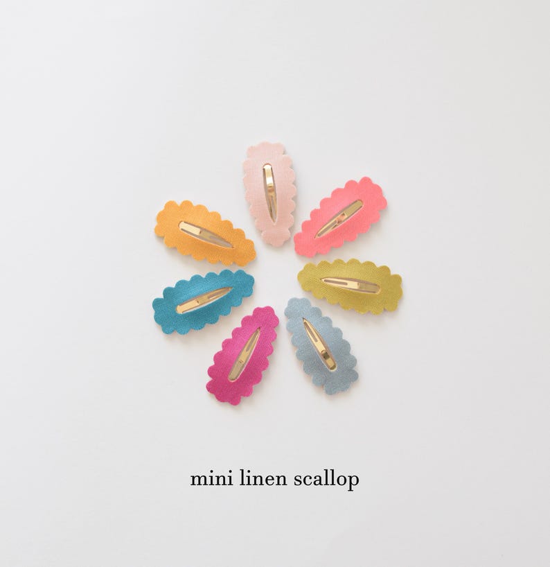 May include: A set of eight mini linen scallop hair clips in various colors, including pink, blue, yellow, and green. The clips are arranged in a circle on a white background.