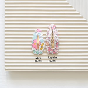 May include: Two floral patterned hair clips, one in pink and one in blue, with gold metal clasps. The smaller clip is labeled "Mini 45mm" and the larger clip is labeled "Regular 55mm".
