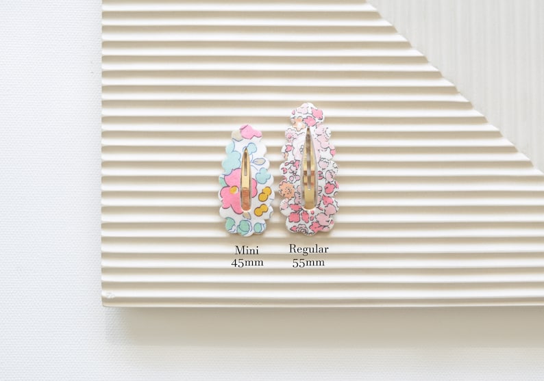 May include: Two floral patterned hair clips, one in pink and one in blue, with gold metal clasps. The smaller clip is labeled "Mini 45mm" and the larger clip is labeled "Regular 55mm".